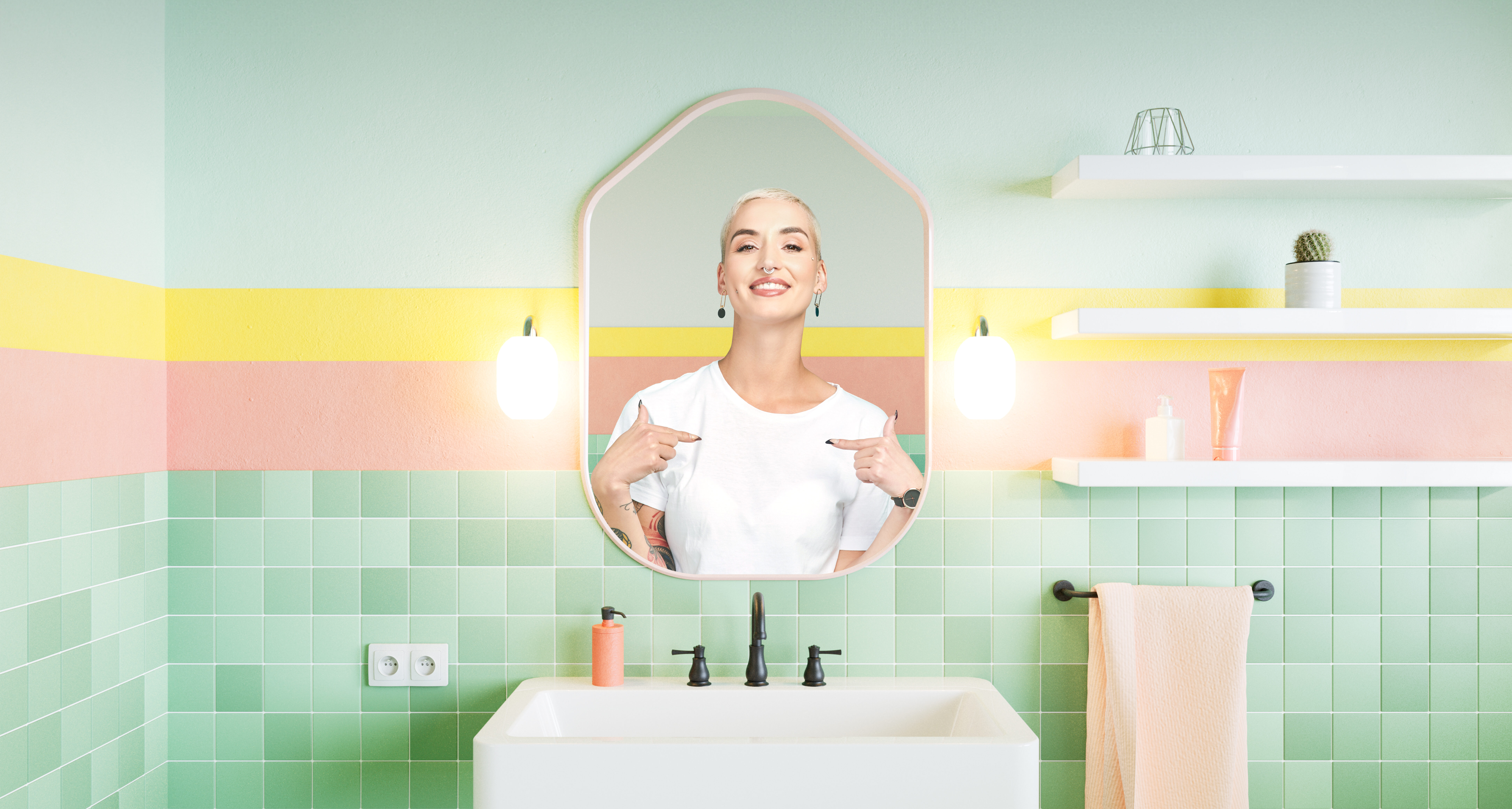 Frau mit weißem T-Shirt lächelt selbstbewusst in einem bunten Badezimmer in den Spiegel und zeigt mit beiden Händen auf sich.