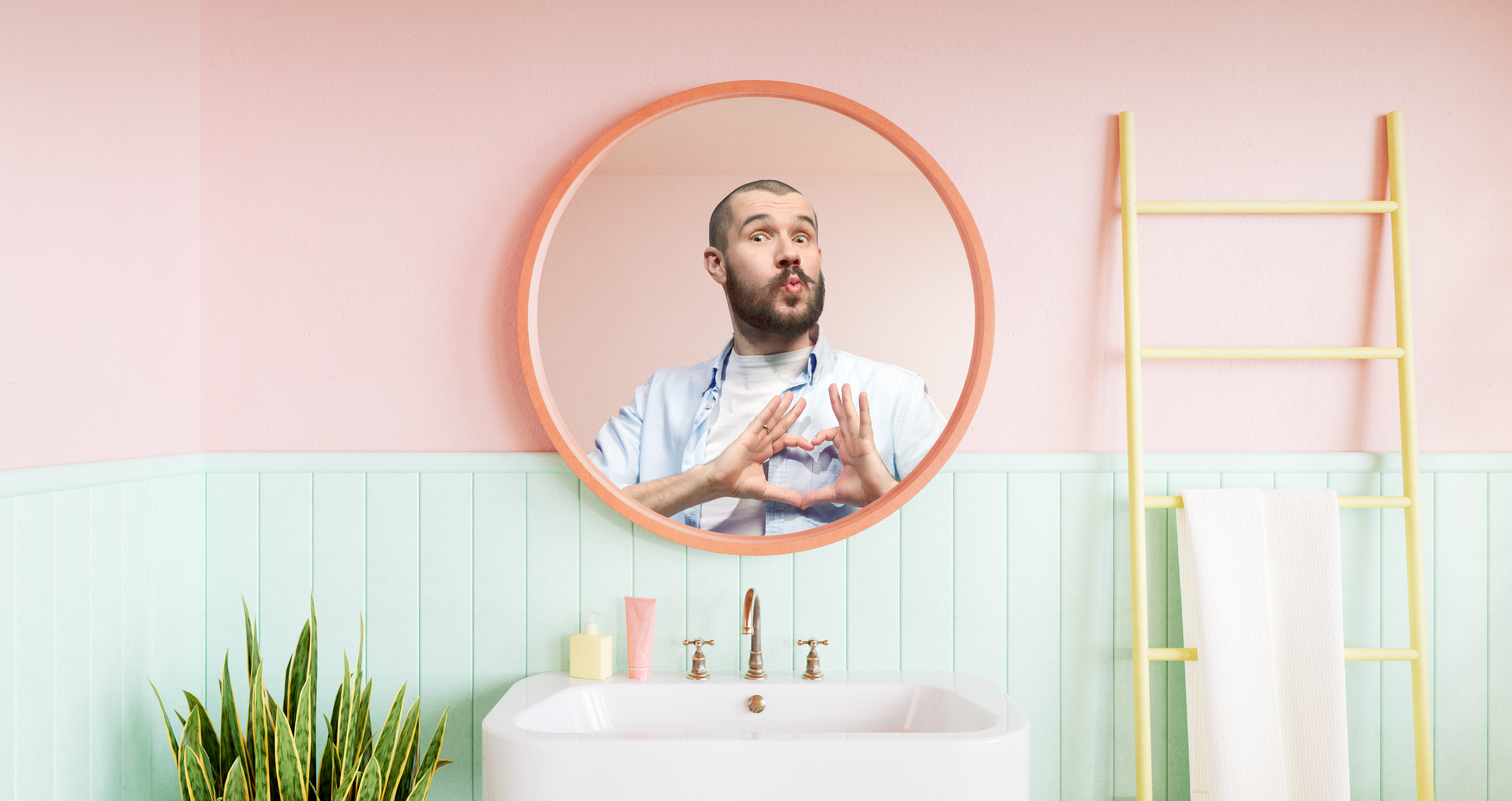 Junger Mann mit rasiertem Kopf formt ein Herz mit den Händen und schaut spielerisch in einem pastellfarbenen Badezimmer in den Spiegel.