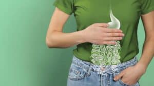 Man sieht den Oberkörper einer Frau, die trägt ein grünes T-Shirt und eine helle Jeans, sie halt ihre Hand vor dem Bauch, darunter ist ein Darm illustriert, der Hintergrund ist grün.