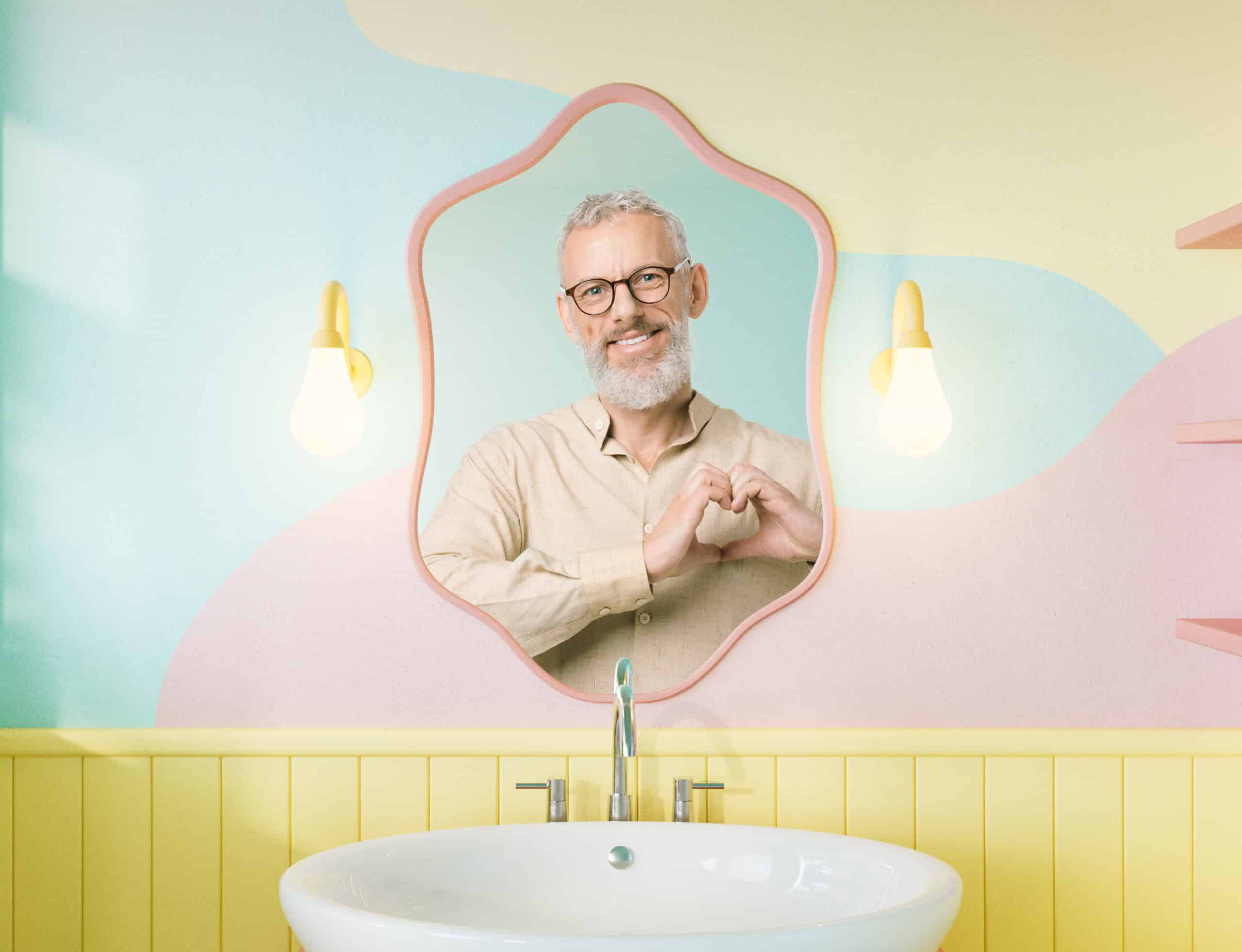 Ősz szakállú, szemüveges idős férfi szív alakot formál a kezével egy pasztellszínű fürdőszoba tükrében.
